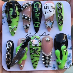 Peut inclure: Un ensemble de dix faux ongles avec une palette de couleurs vert citron et noir, ornés d'éléments 3D tels que des chaînes argentées, des clous métalliques, un couteau miniature et une étoile. Un ongle porte le texte I'M THE SHIT! et un autre Gnarly.