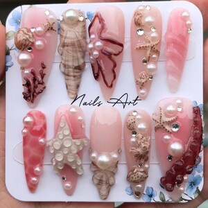 Peut inclure: Dix faux ongles de différentes nuances de rose, ornés de coquillages, de perles et d'étoiles de mer. Certains ongles présentent des motifs coralliens et le texte "Nails Art". Les ongles ont une finition brillante.