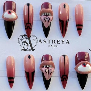 Puede incluir: Un conjunto de diez uñas postizas largas en forma de almendra. Las uñas presentan una variedad de diseños, incluyendo un ojo 3D, una boca y degradados de color negro, burdeos y rosa. Se ve el nombre de la marca "ASTREYA NAILS".