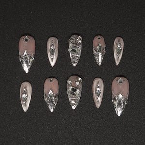 Puede incluir: Un juego de diez uñas postizas en un tono rosa nude suave con forma de almendra puntiaguda. Cada uña presenta detalles de purpurina plateada y pedrería transparente facetada de varios tamaños dispuesta en intrincados patrones geométricos.