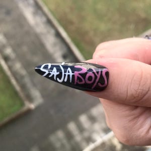 Puede incluir: Una u&ntilde;a postiza negra brillante en forma de almendra con el texto SAJA en blanco y BOYS en rosa, escrito con una fuente manuscrita estilizada en la superficie de la u&ntilde;a. El dise&ntilde;o est&aacute; centrado en la u&ntilde;a, que tiene un acabado suave.