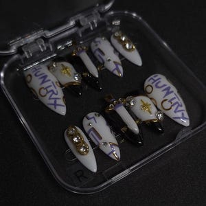 Puede incluir: Un juego de diez u&ntilde;as postizas en forma de estilete en un estuche de pl&aacute;stico transparente. Las u&ntilde;as tienen una base blanca con puntas negras, l&iacute;neas moradas, estrellas doradas, pedrer&iacute;a transparente y el texto HUNTRY escrito en morado.