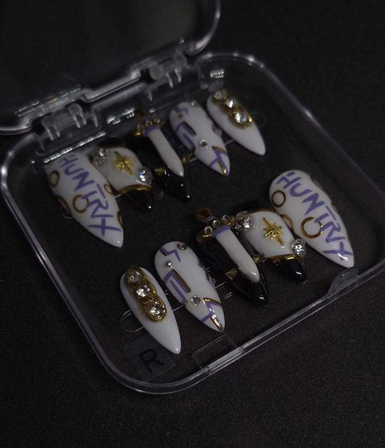 Puede incluir: Un juego de diez u&ntilde;as postizas en forma de estilete en un estuche de pl&aacute;stico transparente. Las u&ntilde;as tienen una base blanca con puntas negras, l&iacute;neas moradas, estrellas doradas, pedrer&iacute;a transparente y el texto HUNTRY escrito en morado.