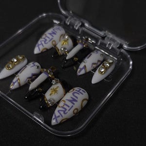 Puede incluir: Un juego de diez u&ntilde;as postizas en forma de estilete en un estuche de pl&aacute;stico transparente. Las u&ntilde;as presentan una paleta de colores blanco y negro con detalles dorados, pedrer&iacute;a, peque&ntilde;os dijes de espada y la palabra HUNTRX en morado.