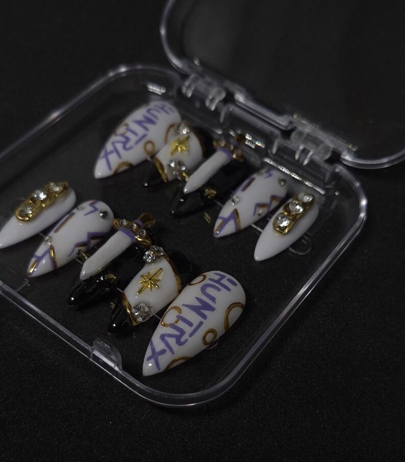 Puede incluir: Un juego de diez u&ntilde;as postizas en forma de estilete en un estuche de pl&aacute;stico transparente. Las u&ntilde;as presentan una paleta de colores blanco y negro con detalles dorados, pedrer&iacute;a, peque&ntilde;os dijes de espada y la palabra HUNTRX en morado.