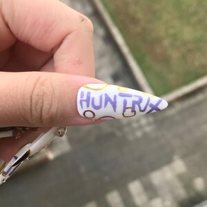 Puede incluir: Una u&ntilde;a postiza blanca en forma de estilete con el texto "HUNTRX" escrito a mano en color morado en el centro. El dise&ntilde;o de la u&ntilde;a incluye contornos circulares dorados y marrones dispersos, creando un patr&oacute;n decorativo sobre la base blanca.