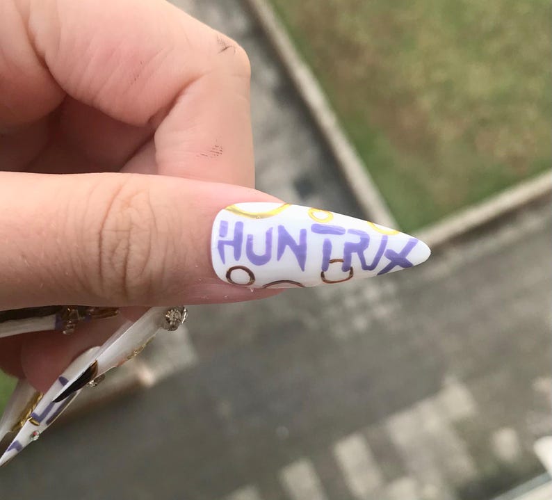 Puede incluir: Una u&ntilde;a postiza blanca en forma de estilete con el texto "HUNTRX" escrito a mano en color morado en el centro. El dise&ntilde;o de la u&ntilde;a incluye contornos circulares dorados y marrones dispersos, creando un patr&oacute;n decorativo sobre la base blanca.