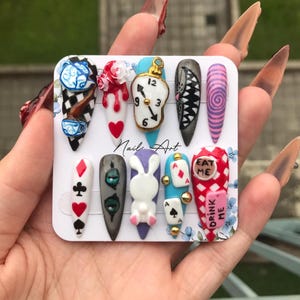 Puede incluir: Un conjunto de diez uñas acrílicas largas y puntiagudas con temática de Alicia en el país de las maravillas. Los diseños incluyen un reloj, un conejo, naipes y las palabras "Eat Me" y "Drink Me". Las uñas están pintadas en varios colores, incluyendo blanco, rojo, azul y negro.