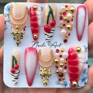 May include: A set of artificial nails with various designs. Some nails feature cherries, gold accents, and zebra stripes. Others have red and white patterns, gold crosses, and pearl embellishments. The text "Nails Art" is visible.