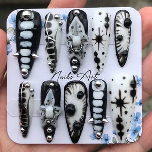 Puede incluir: Un conjunto de diez uñas artificiales con un diseño en blanco y negro. Las uñas presentan varios patrones, incluyendo lunares, rayas y detalles florales. Algunas uñas tienen tachuelas y púas plateadas. El texto "Nails Art" es visible.
