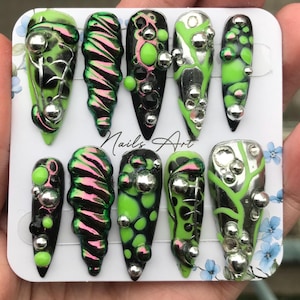 Peut inclure: Un ensemble de dix faux ongles avec divers motifs. Les ongles présentent une combinaison de noir, de vert et d'argent. Certains ont un motif tourbillonnant, d'autres un motif de bulles ou de toile d'araignée. Le texte "Nails Art" est visible.