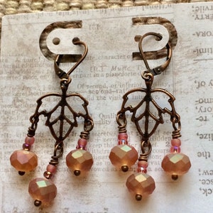 May include: A pair of copper-toned earrings with a leaf design and dangling amber-colored glass beads. The earrings are hanging from a white card with text.