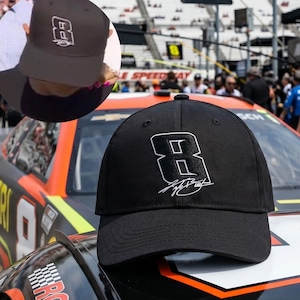 May include: Black baseball cap featuring a white number 8 and a signature. The cap is displayed on the hood of a race car. The car is black, orange, and green. The background shows a racetrack and spectators.