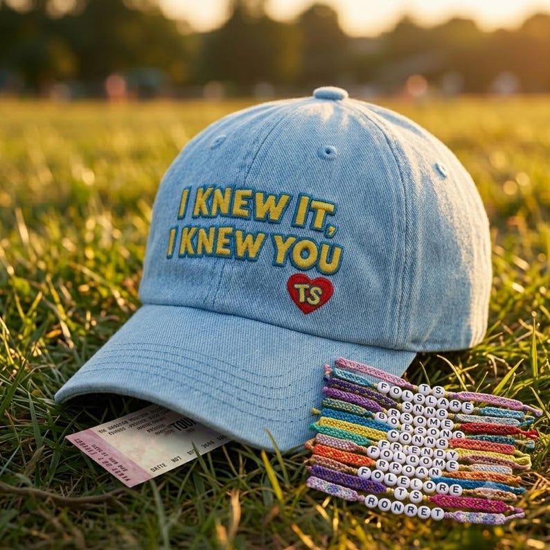K&ouml;nnte beinhalten: Eine hellblaue Baseballkappe aus Denim mit gelbem Sticktext, der I KNEW IT, I KNEW YOU lautet, &uuml;ber einem roten Herz mit den Buchstaben TS. Neben der Kappe liegen mehrere bunte Freundschaftsarmb&auml;nder mit wei&szlig;en Buchstabenperlen.