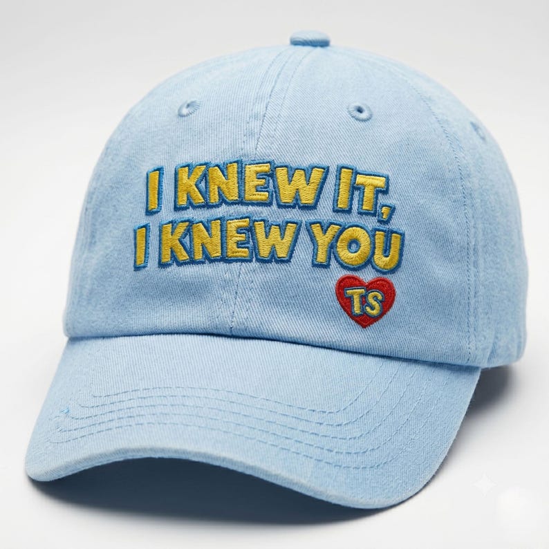 K&ouml;nnte beinhalten: Eine hellblaue Baseballkappe mit dem gestickten gelben Text I KNEW IT, I KNEW YOU und einem roten Herzsymbol, das die gelben Buchstaben TS enth&auml;lt. Die Kappe hat einen gebogenen Schirm, einen Knopf oben und gen&auml;hte &Ouml;sen zur Bel&uuml;ftung.