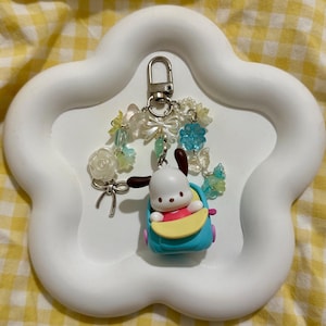 May include: A white, cloud-shaped tray holds a keychain featuring a cartoon dog in a blue car. The keychain has a silver clasp and is adorned with clear and blue beads, bows, and flowers. The background is a yellow and white checkered pattern.