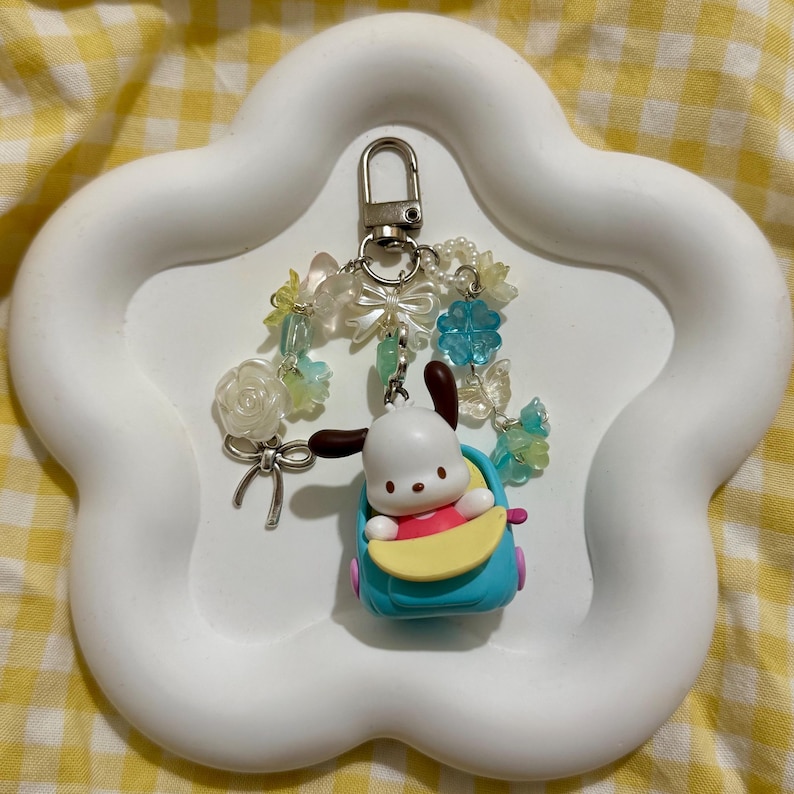 May include: A white, cloud-shaped tray holds a keychain featuring a cartoon dog in a blue car. The keychain has a silver clasp and is adorned with clear and blue beads, bows, and flowers. The background is a yellow and white checkered pattern.