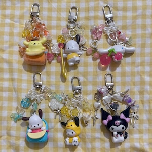 May include: Six colorful keychains with cartoon character charms. Each keychain features a silver clasp, decorative beads, bows, and a unique character figurine. The characters include a yellow dog, a white dog, a white bunny, and a black cat.