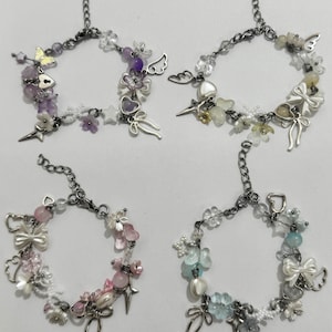 May include: Four decorative bracelets with silver chains and charms. Each bracelet has a unique design with beads, flowers, bows, and star accents in various colours, including purple, pink, white, and blue. The bracelets are designed for a feminine aesthetic.