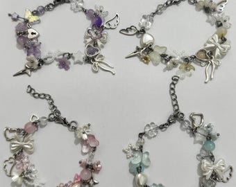 May include: Four delicate bracelets with silver-tone chains and charms. Each bracelet features a unique design with beads, flowers, bows, and star accents in various colors, including purple, pink, white, and blue. The bracelets are designed for a feminine aesthetic.