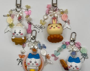 May include: Four decorative keychains featuring cartoon character figures, including a white creature in a brown bowl, a yellow rabbit-like figure, a white cat-like figure in a pink bowl, and a white cat-like figure holding a brown cookie, all with beads and charms.
