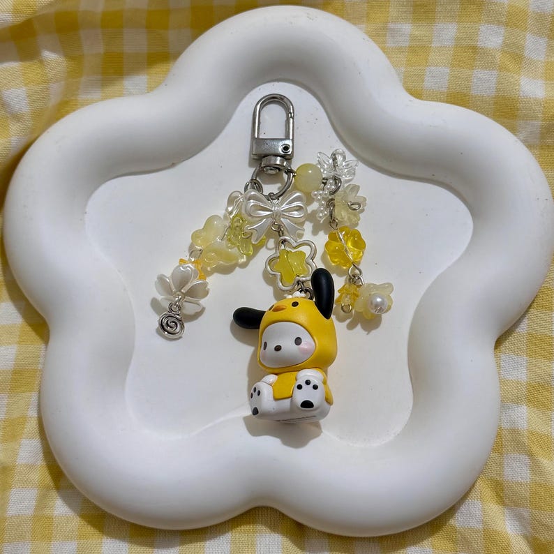 May include: A yellow and white character keychain with a silver clasp, adorned with yellow and white floral and bow charms. The character is wearing a yellow outfit with black accents, sitting on a white, cloud-shaped tray.