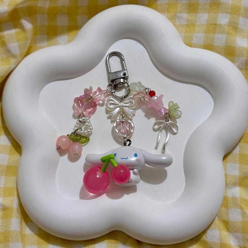 May include: A white keychain featuring a cartoon character with a cherry design. The keychain has a silver clasp and is adorned with pink and clear beads, bows, and flowers. The keychain is displayed on a white, cloud-shaped tray.