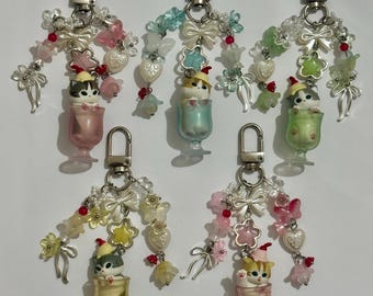 May include: Five keychains featuring small cat figurines in colorful, translucent cups. Each keychain has a silver clasp and is adorned with decorative beads, bows, and flower-shaped charms in various pastel colors. The cats wear tiny Santa hats.