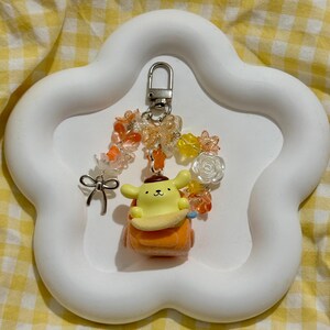 May include: A yellow and orange cartoon character keychain with a silver clasp, adorned with orange and yellow beads, a silver bow, and flower accents. The keychain is displayed on a white, cloud-shaped tray.