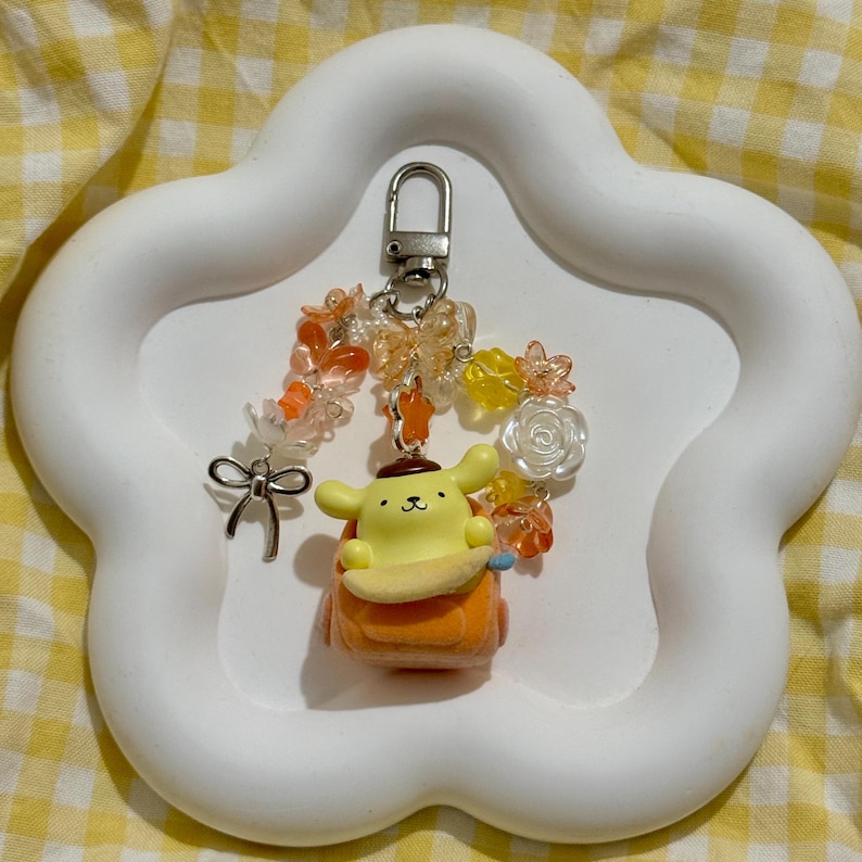 May include: A yellow and orange cartoon character keychain with a silver clasp, adorned with orange and yellow beads, a silver bow, and flower accents. The keychain is displayed on a white, cloud-shaped tray.