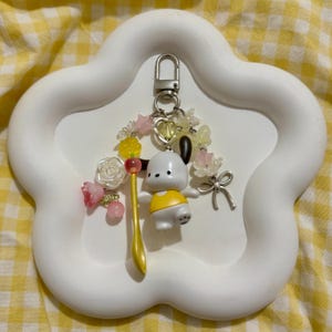 May include: A white and yellow keychain featuring a cartoon dog character with a yellow shirt, a golden spoon, and decorative floral accents. The keychain is attached to a silver clasp and is set against a white, cloud-shaped tray.