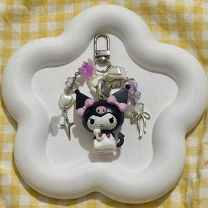 May include: A black and white character keychain with pink accents, a devil tail, and a skull on its forehead. The keychain is adorned with beads, charms, and a silver clasp. It is displayed on a white, cloud-shaped tray.