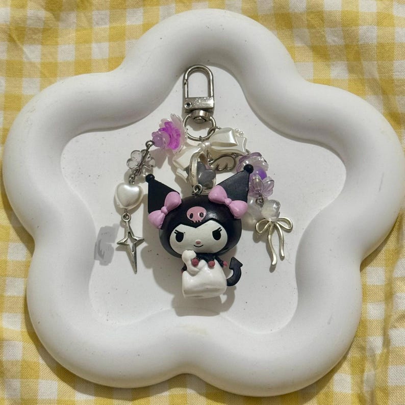 May include: A black and white character keychain with pink accents, a devil tail, and a skull on its forehead. The keychain is adorned with beads, charms, and a silver clasp. It is displayed on a white, cloud-shaped tray.