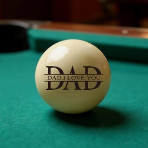 May include: A cream-colored billiard ball with the word "DAD" and "DAD, I LOVE YOU" printed in dark brown. The ball is resting on a green felt pool table.