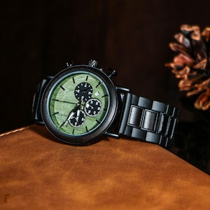 Puede incluir: Un reloj de pulsera de madera con esfera verde y detalles en negro. El reloj tiene tres subesferas y una correa de madera negra. El reloj se muestra sobre una superficie marr&oacute;n.