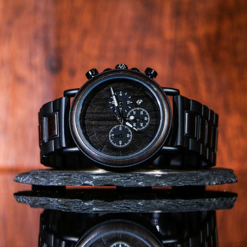 Puede incluir: Un reloj de madera negro con esfera de grano de madera oscura y correa de metal negro. El reloj tiene tres subesferas y un bisel negro. El reloj se muestra sobre una superficie de piedra gris oscura.