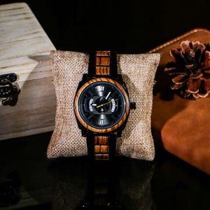 Puede incluir: Un reloj de pulsera de madera con esfera negra y correa de madera marr&oacute;n. El reloj se exhibe sobre un coj&iacute;n peque&ntilde;o y cuadrado de color beige. El reloj tiene un bisel negro y un segundero amarillo. Un accesorio elegante.