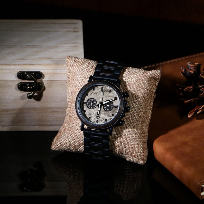 Puede incluir: Un reloj de pulsera negro con esfera de madera y correa de metal negro descansa sobre un coj&iacute;n de arpillera. La esfera del reloj presenta un dise&ntilde;o texturizado de color claro con tres esferas m&aacute;s peque&ntilde;as. Una caja de madera y un cuaderno de cuero est&aacute;n en el fondo.