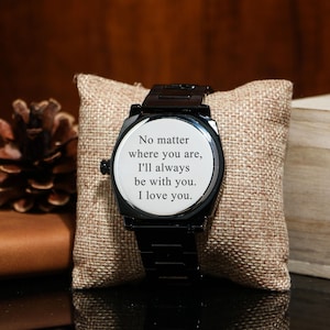 Puede incluir: Reloj de pulsera negro con esfera blanca que muestra el texto: "No importa d&oacute;nde est&eacute;s, siempre estar&eacute; contigo. Te amo". El reloj est&aacute; sobre una almohada de arpillera.
