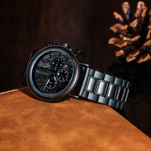 Puede incluir: Un reloj cron&oacute;grafo con esfera de madera y una correa de metal negro. La esfera del reloj presenta tres subesferas y un patr&oacute;n de vetas de madera oscura. El reloj est&aacute; sobre una superficie de cuero marr&oacute;n.