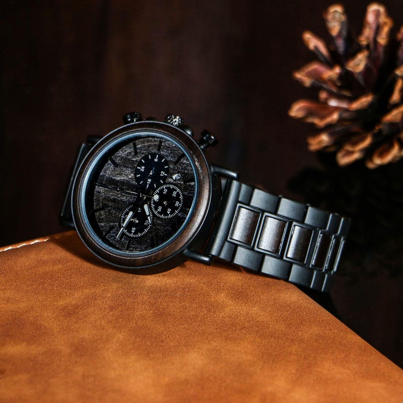 Puede incluir: Un reloj cron&oacute;grafo con esfera de madera y una correa de metal negro. La esfera del reloj presenta tres subesferas y un patr&oacute;n de vetas de madera oscura. El reloj est&aacute; sobre una superficie de cuero marr&oacute;n.
