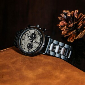Puede incluir: Un reloj de pulsera de madera con esfera gris oscuro y detalles en negro. El reloj presenta una esfera gris texturizada con dos esferas m&aacute;s peque&ntilde;as y una correa negra. El reloj se muestra sobre una superficie de cuero marr&oacute;n.