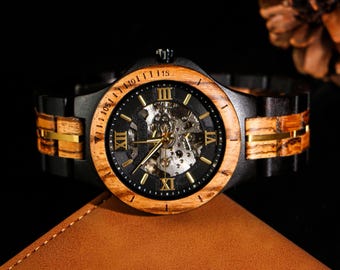 Puede incluir: Un reloj de pulsera de madera con una correa de cuero negro y un movimiento mecánico visible. La esfera del reloj presenta números romanos y agujas doradas. El reloj se muestra sobre una superficie marrón.
