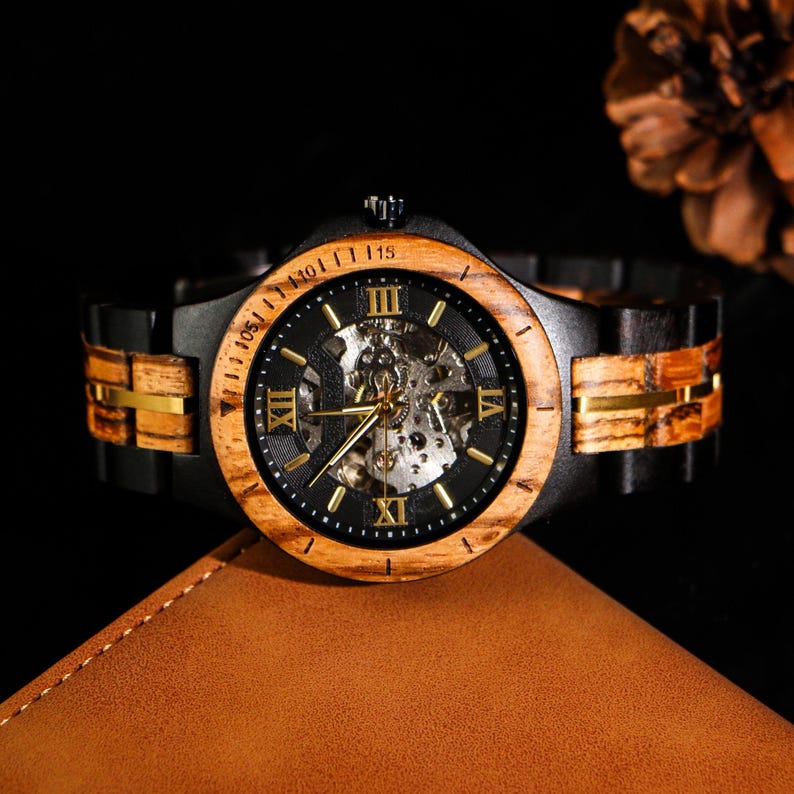 May include: A wooden wristwatch with a black face and gold hands. The watch has a wooden bezel and band with gold accents. The watch is displayed on a brown leather surface.