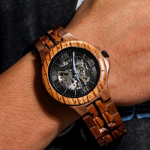 May include: Wooden wristwatch with a dark face and visible inner workings. The watch has a light brown wooden bezel and band. The watch hands are blue. The watch face features Roman numerals.