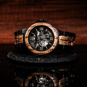 May include: A wooden wristwatch with a black face and a visible mechanical movement. The watch has a light brown wooden bezel and a black band with wooden accents. The watch is displayed on a black surface with a reflective surface below.