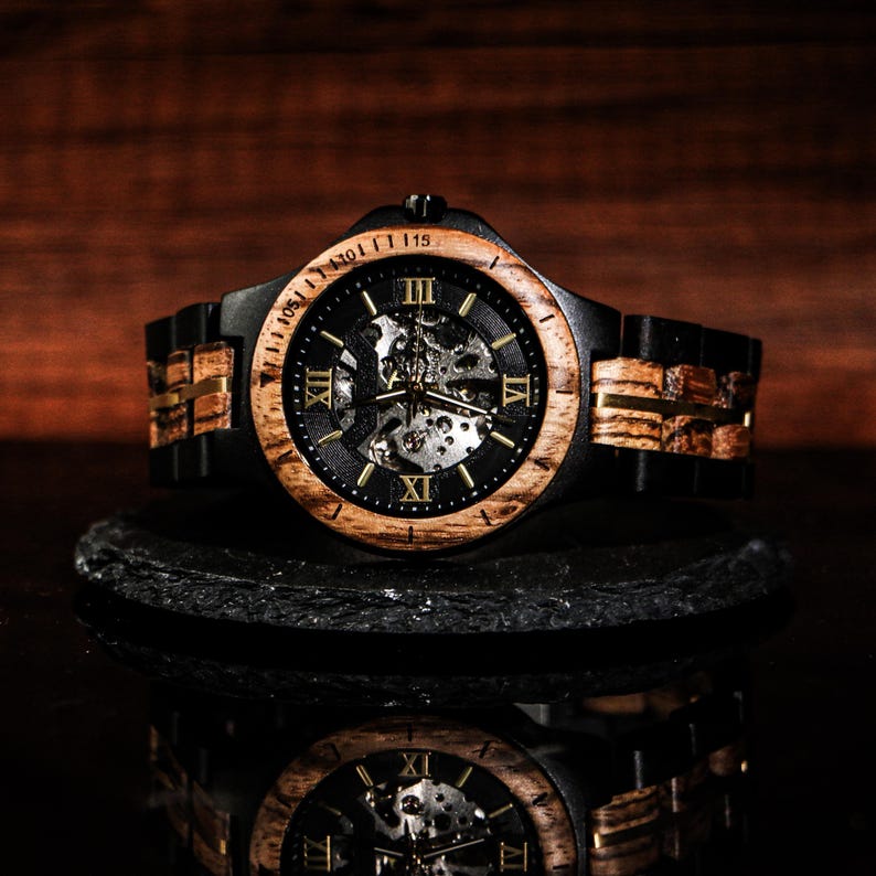 May include: A wooden wristwatch with a black face and a visible mechanical movement. The watch has a light brown wooden bezel and a black band with wooden accents. The watch is displayed on a black surface with a reflective surface below.