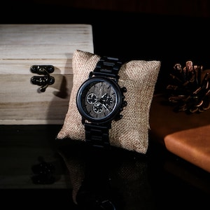 Puede incluir: Un reloj de pulsera negro con una esfera de madera y tres subesferas descansa sobre una almohada de arpillera. El reloj tiene una correa de metal negro y una esfera de madera oscura. Una peque&ntilde;a caja de madera con un cierre de metal est&aacute; en el fondo.