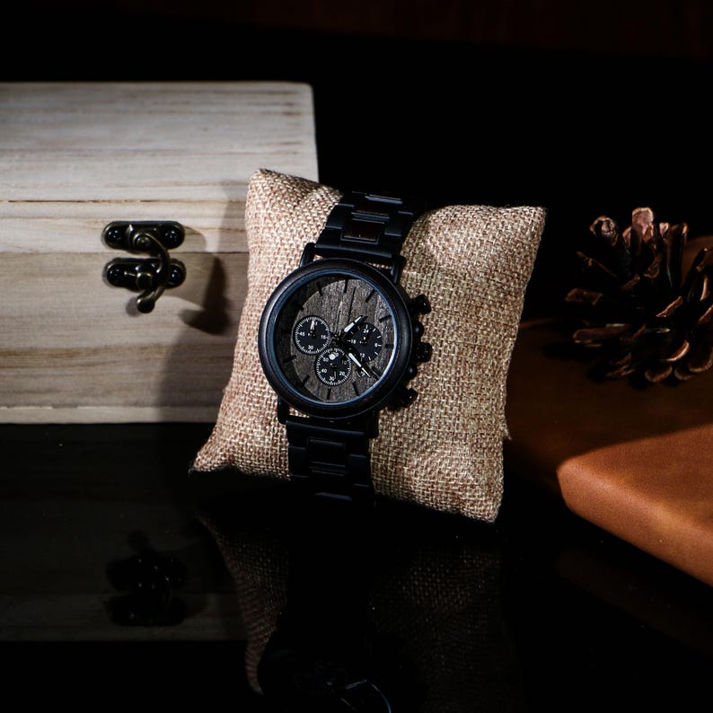 Puede incluir: Un reloj de pulsera negro con una esfera de madera y tres subesferas descansa sobre una almohada de arpillera. El reloj tiene una correa de metal negro y una esfera de madera oscura. Una peque&ntilde;a caja de madera con un cierre de metal est&aacute; en el fondo.