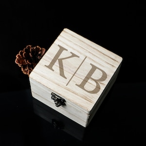 May include: A light-coloured wooden box with the engraved initials "KB" on the lid. The box has a small, dark metal clasp and is next to a brown pine cone. The background is black.